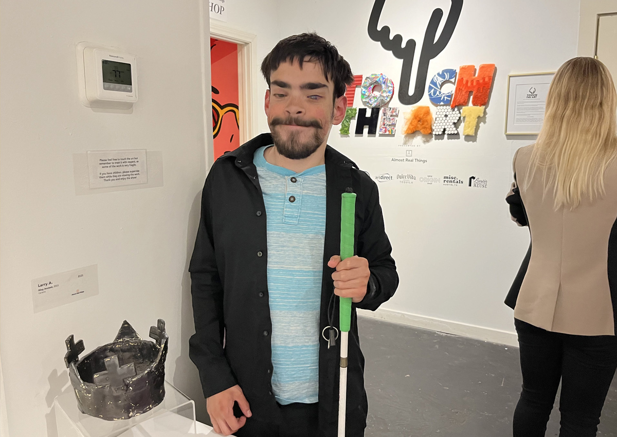 An adolescent in an art gallery stands next to a ceramic crown with fiber art sculptures, weavings, and pottery also on display.