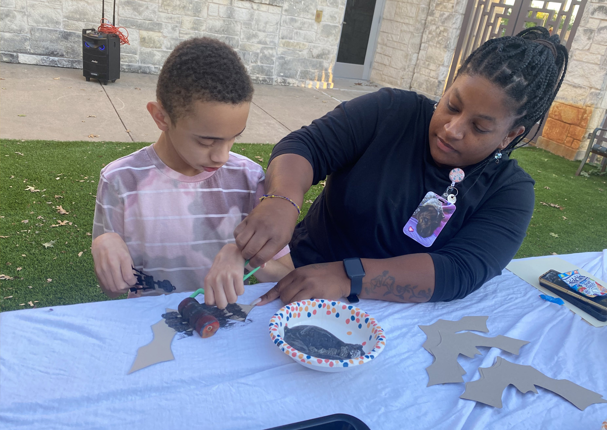 An adult seated next to an adolescent at a picnic table outdoors helps the child use a paint roller to paint a paper bat black.