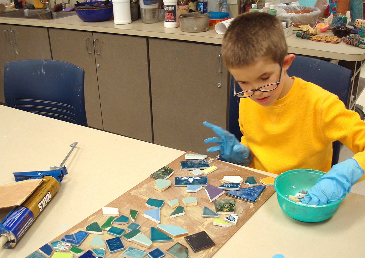 An adolescent wearing gloves glues pieces of different colored tiles onto a rectangular board to make a mosaic.