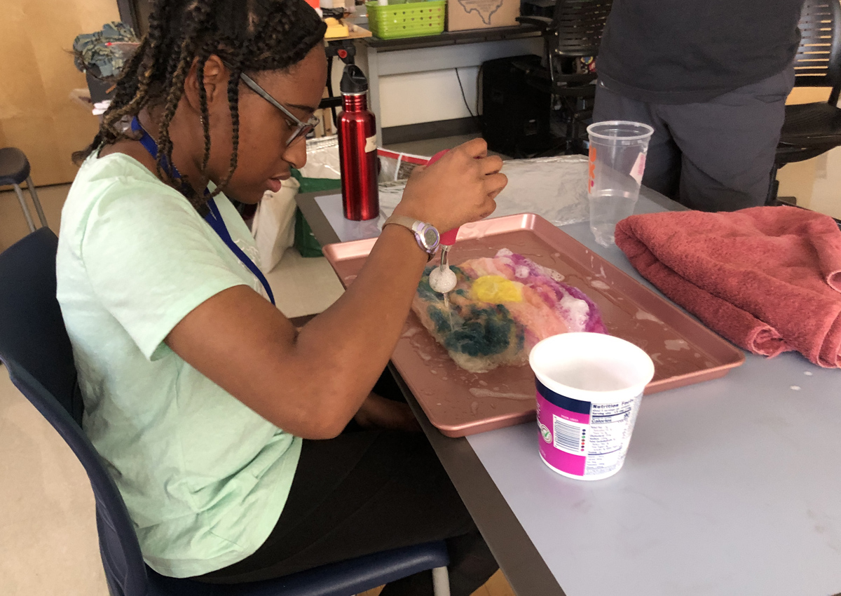 An adolescent drips a thick liquid from a tool onto a square, colorful piece of felt on a tray in an art classroom.