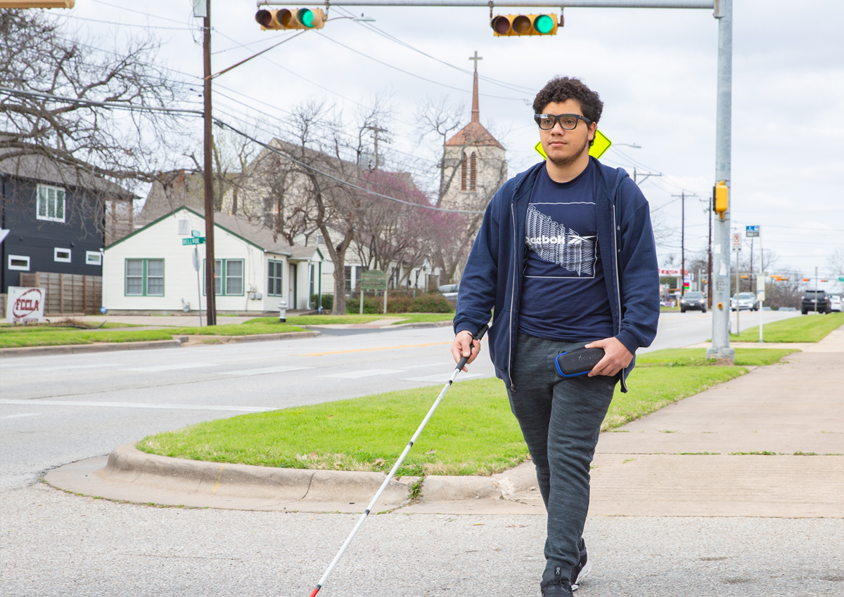 An adolescent wearing Envision glasses uses a cane in his right hand when crossing the street.