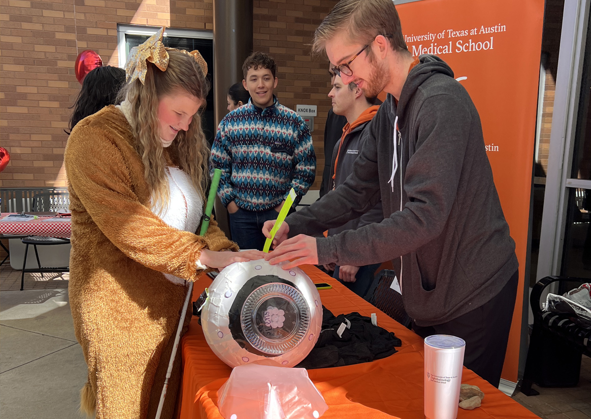 An adult helps an adolescent in a wildcat mascot suit assemble a jumbo model of an eyeball at a Dell Medical school booth.