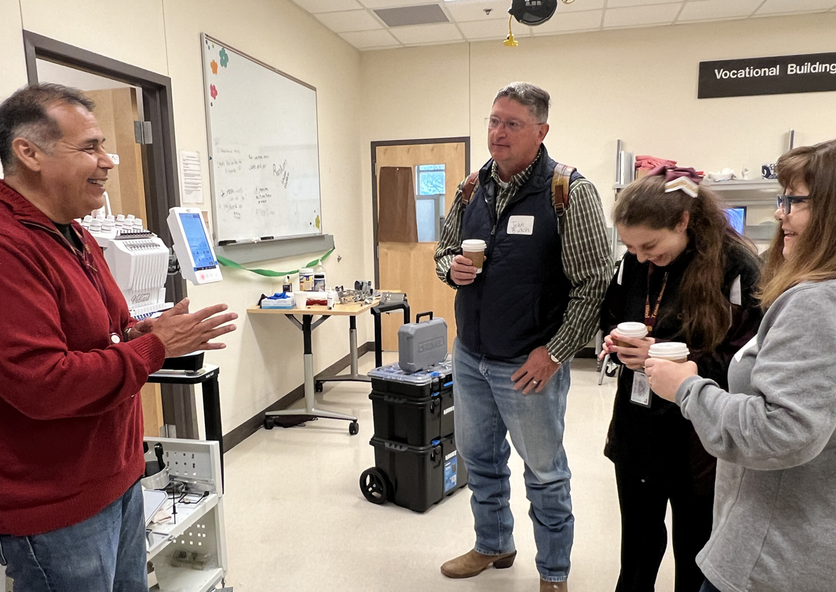 An adult standing next to an embroidery machine talks to an adolescent and their parents in a room labeled 