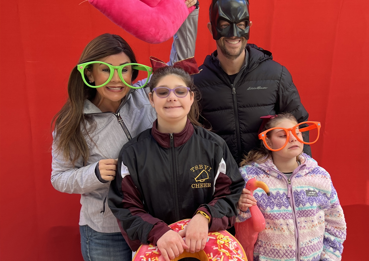 A family of four pose with photo booth props (large glasses, jumbo stuffed donut, and batman mask).