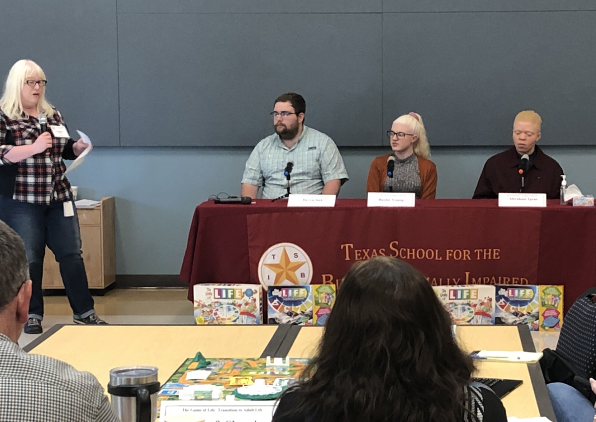 Workshop attendees look at a student panel sitting behind a table with a TSBVI table cloth. A woman stands to the side holding a microphone and paper, asking the panel a question.