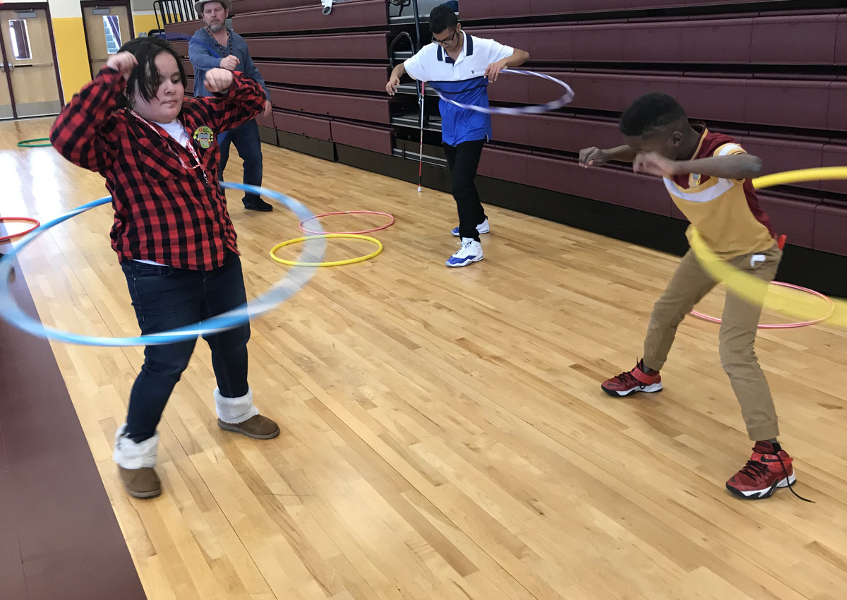 Students and an adult hula hoop in a gymnasium.