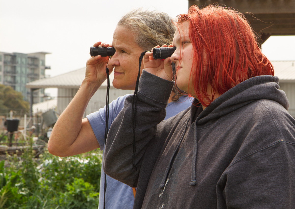 An adult and adolescent hold monoculars while looking at distant objects outside in a community garden.