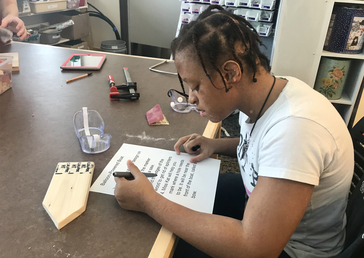 an adolescent sits at a table reading a large-print list with instructions about how to make a balloon-powered boat. Wood and tools are on the table.