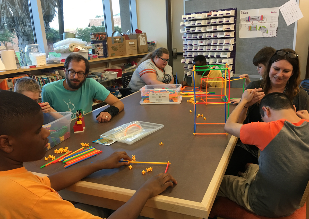Five adolescents and three staff sit at a large table in a library building with tools and plastic straws.