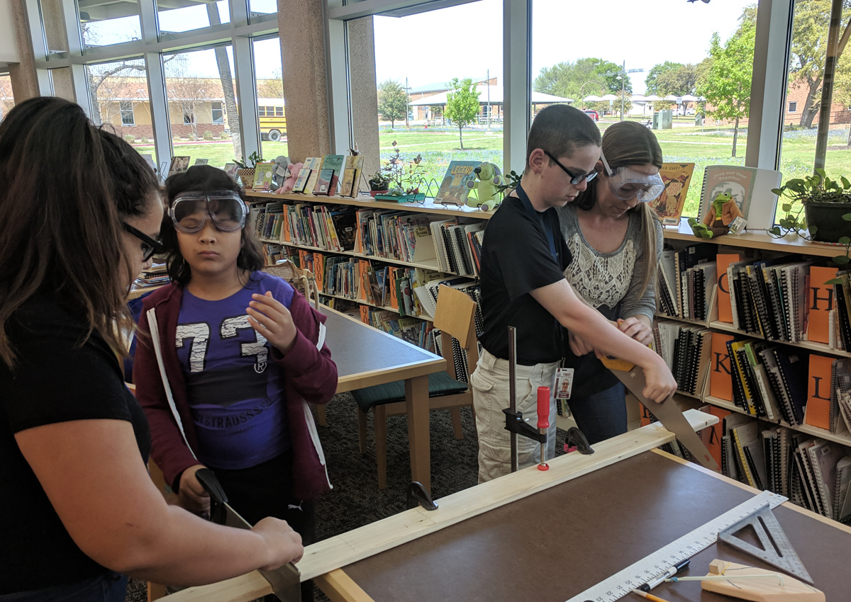 Two adolescents saw wood with the help of two adults in a library.