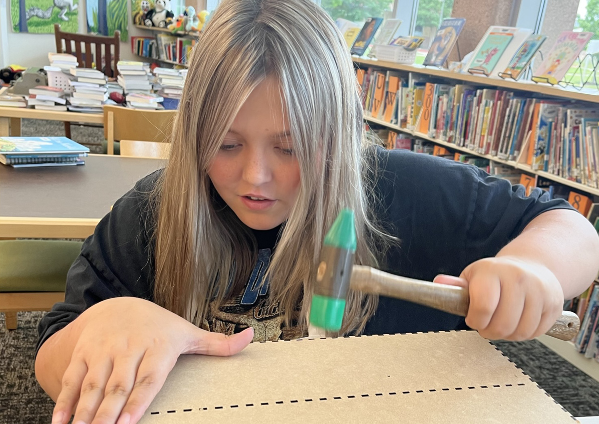 An adolescent hammers pieces of cardboard together into a box that were constructed using a laser cutter.