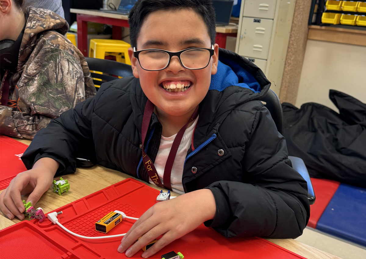An adolescent connects electronic building blocks that snap together with magnets while seated at a table in a classroom.