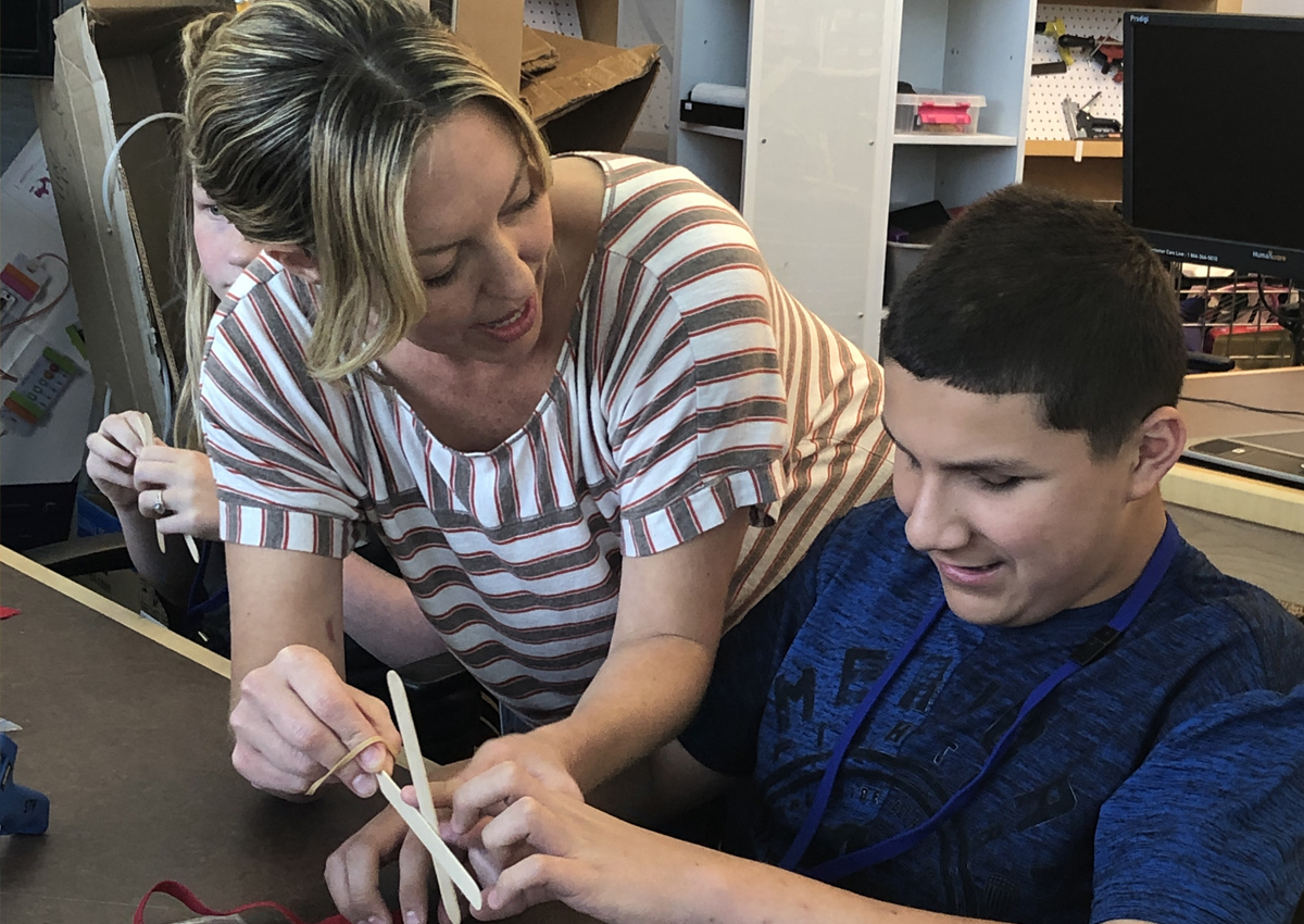 An adolescent builds with popsicle sticks while an adult helps him hold them together using a rubber band.