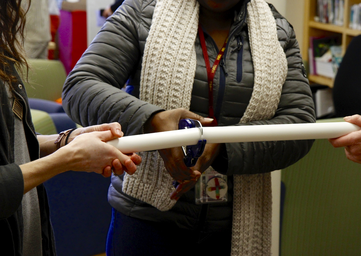 An adult uses clippers to cut a long PVC pipe held by two other adults.