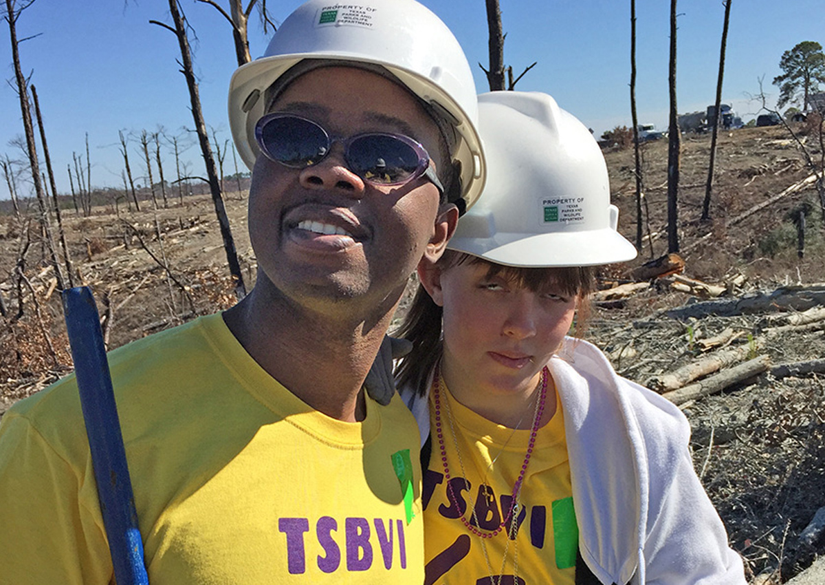Two adolescents wearing hard hats carry shovels in a barren field with burnt tree stumps.
