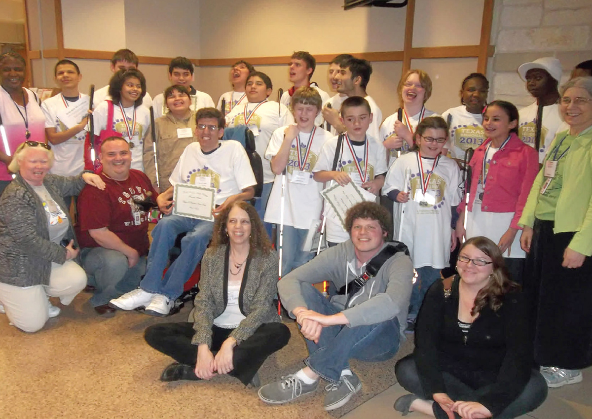A large group of adolescents wearing matching white Braille Challenge t-shirts, holding certificates, and wearing medals and adults stand together.