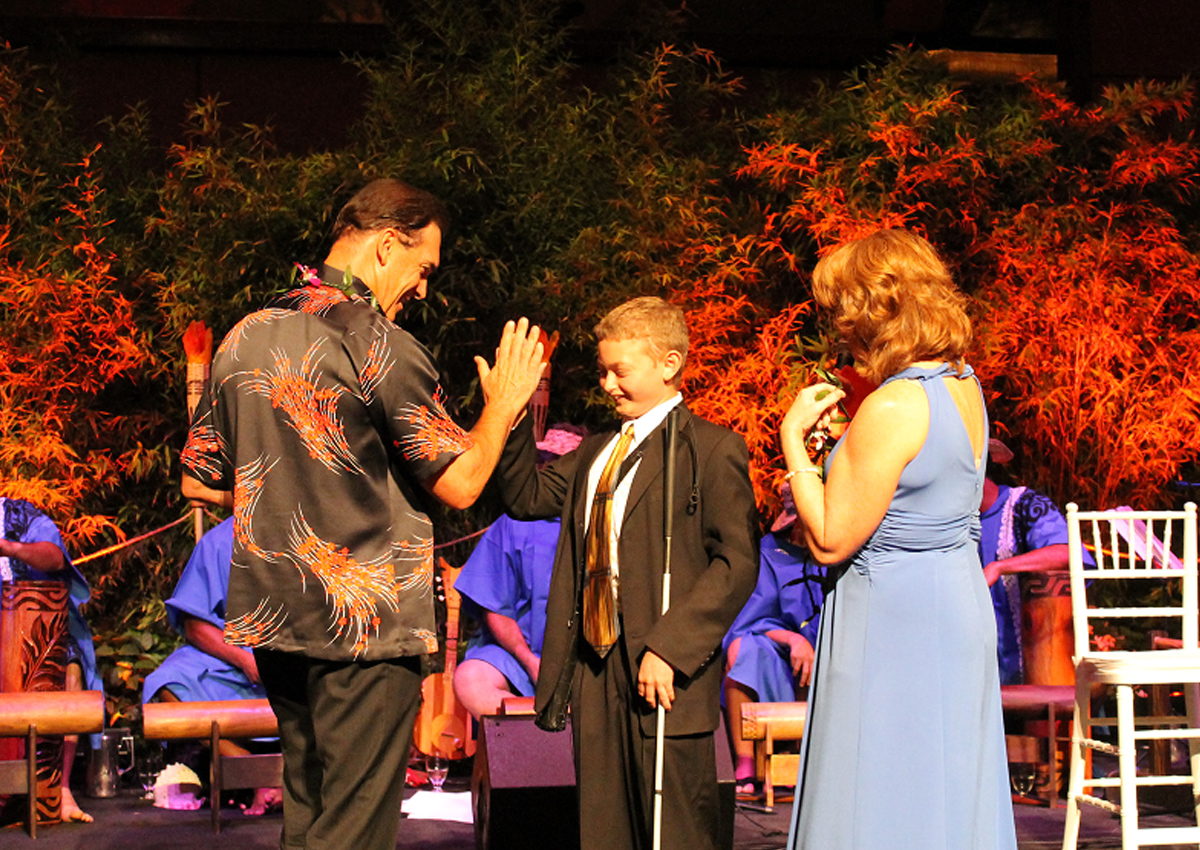An adolescent in a suit high-fives actor Patrick Warburton on a stage with performers.