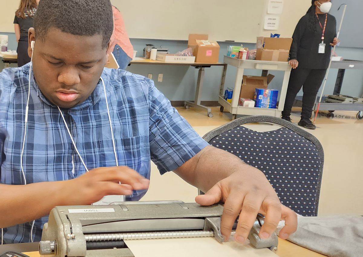 An adolescent wearing earbuds checks Braille he wrote on a Perkins Brailler.