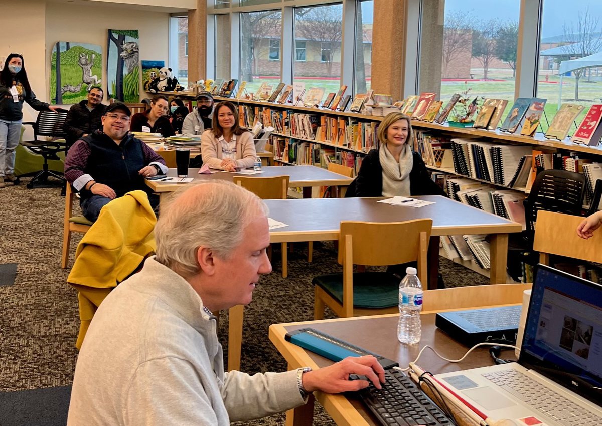 An adult sits at a table in a library presenting on 
