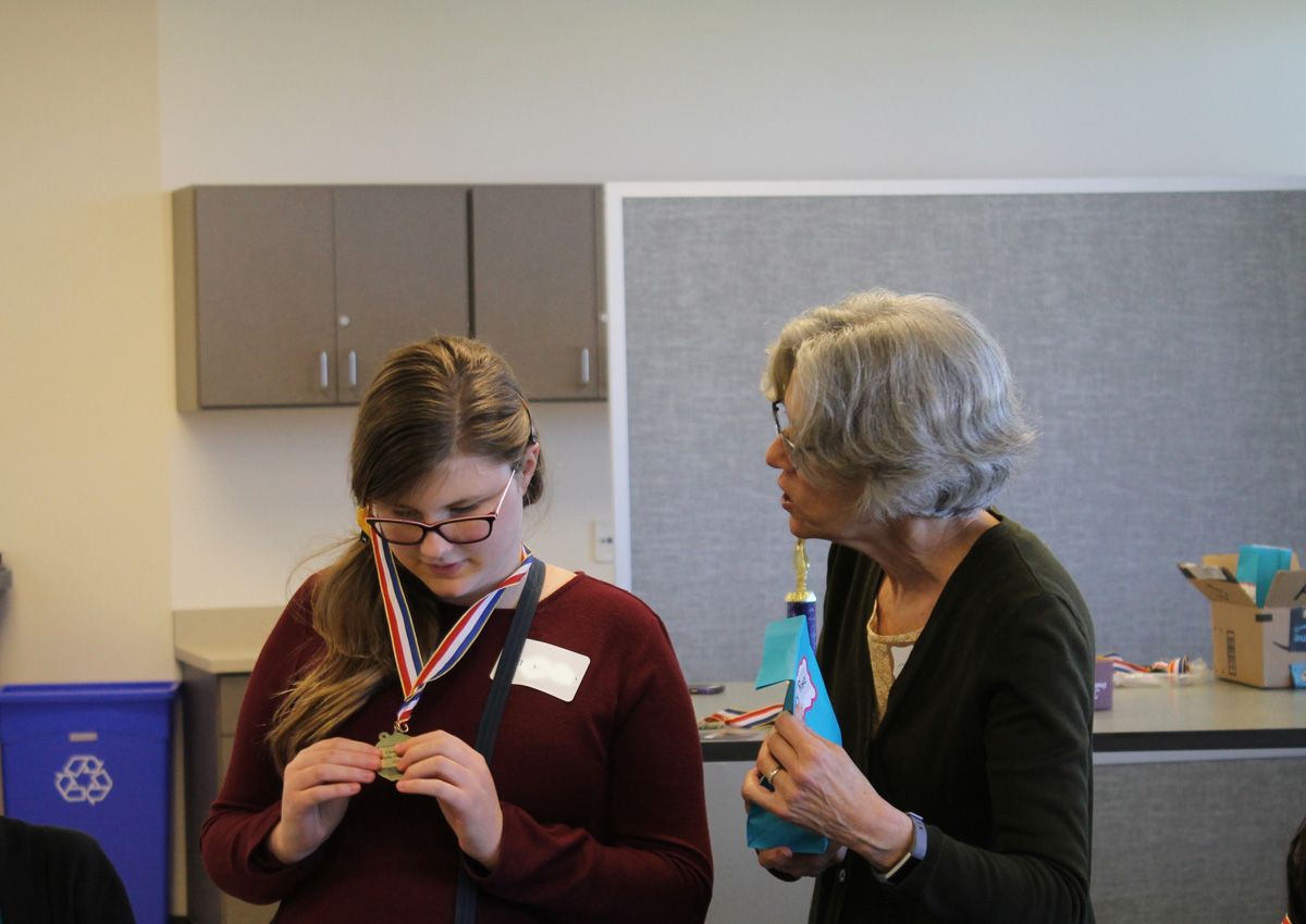 An adolescent looks at a medal around her neck while an adult stands nearby.