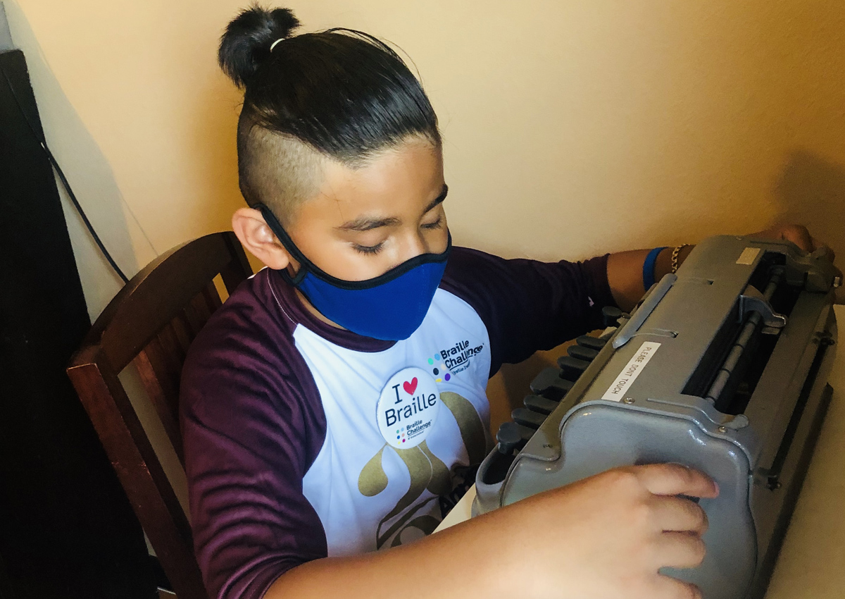 An adolescent uses a Perkins brailler while wearing a Braille Challenge face mask, a 2020 Braille Challenge Finals shirt and a button that reads 