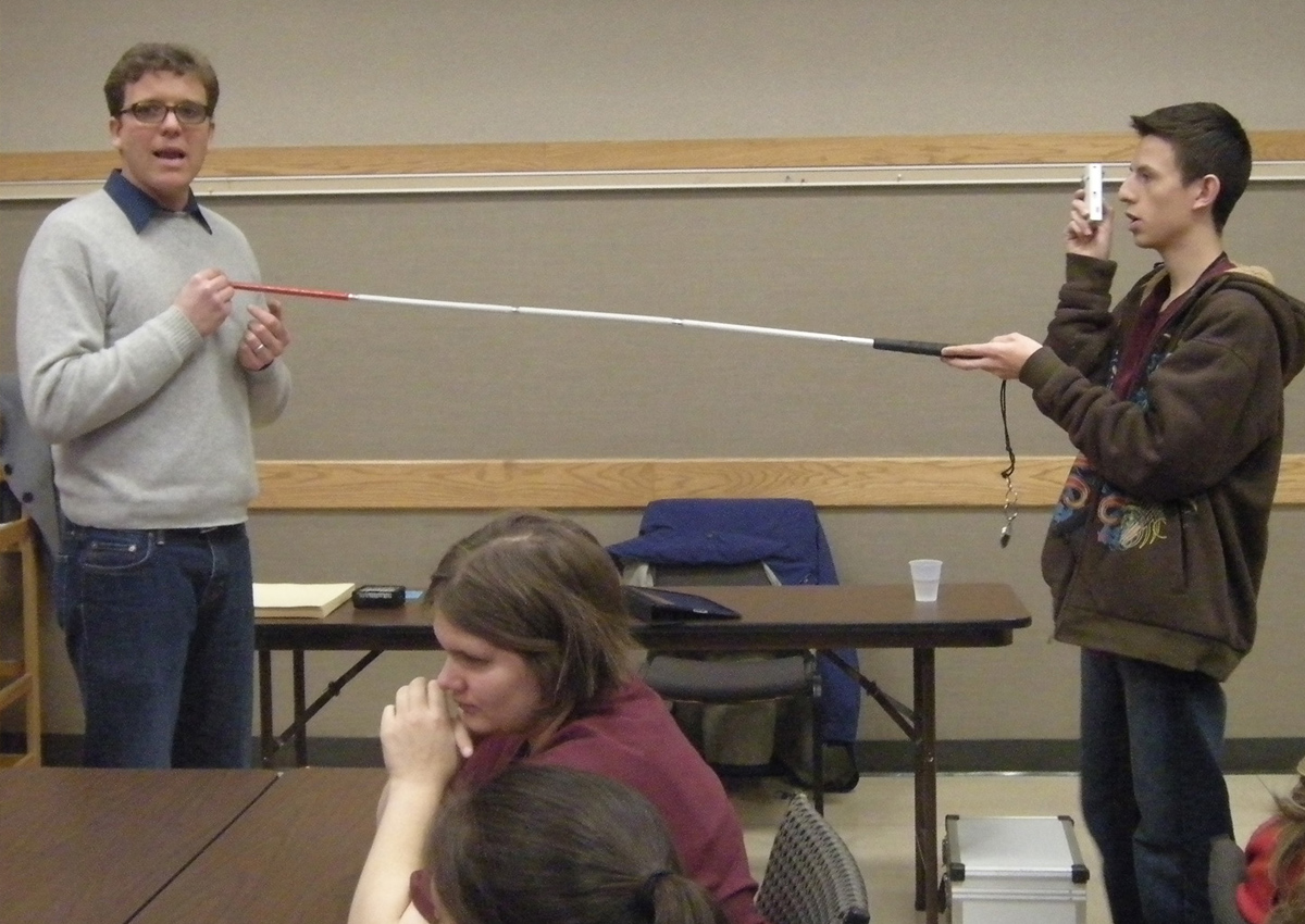 An adolescent and an adult hold a long white cane between them while the adolescent uses a handheld digital video camera to record.