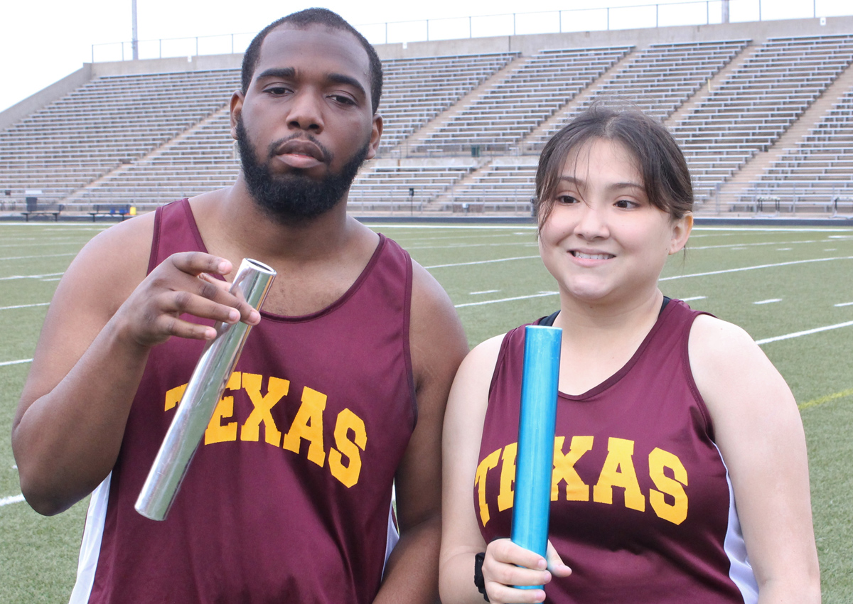 Two adolescents in Texas track jersey's hold up batons while standing in the middle of an outdoor stadium.