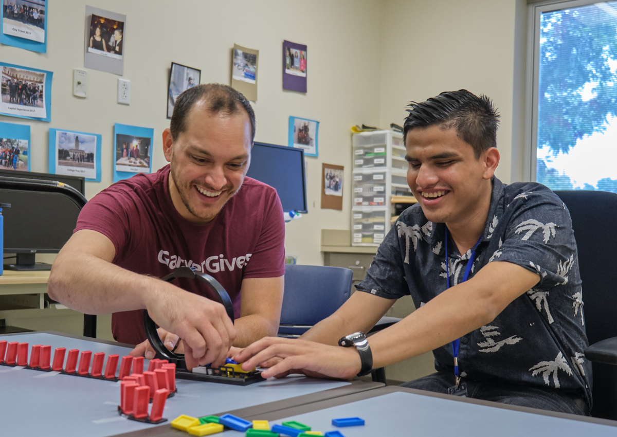 An adult and adolescent explore a hot wheels track together. Dominoes set up in a row on a track are on the table.