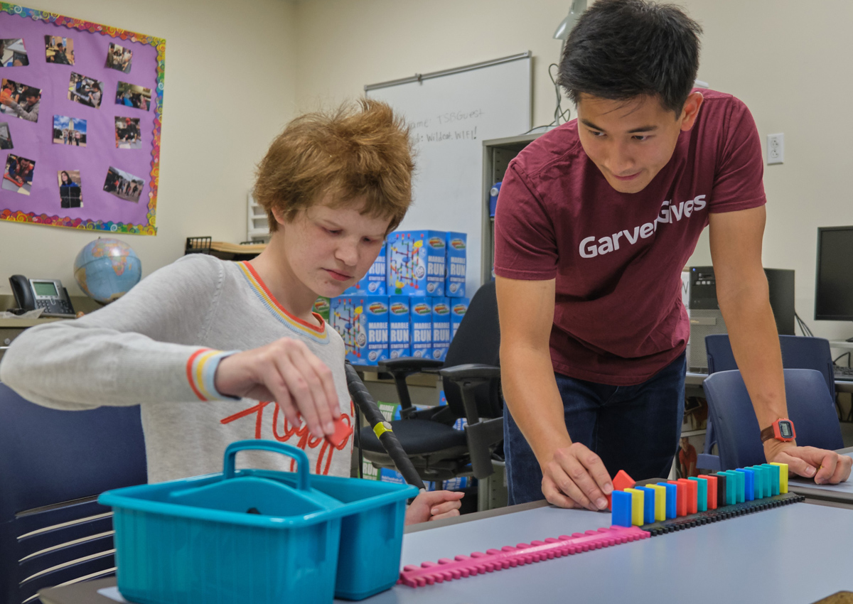 An adolescent seated at a table lines up dominoes alongside a track with an adult's help.