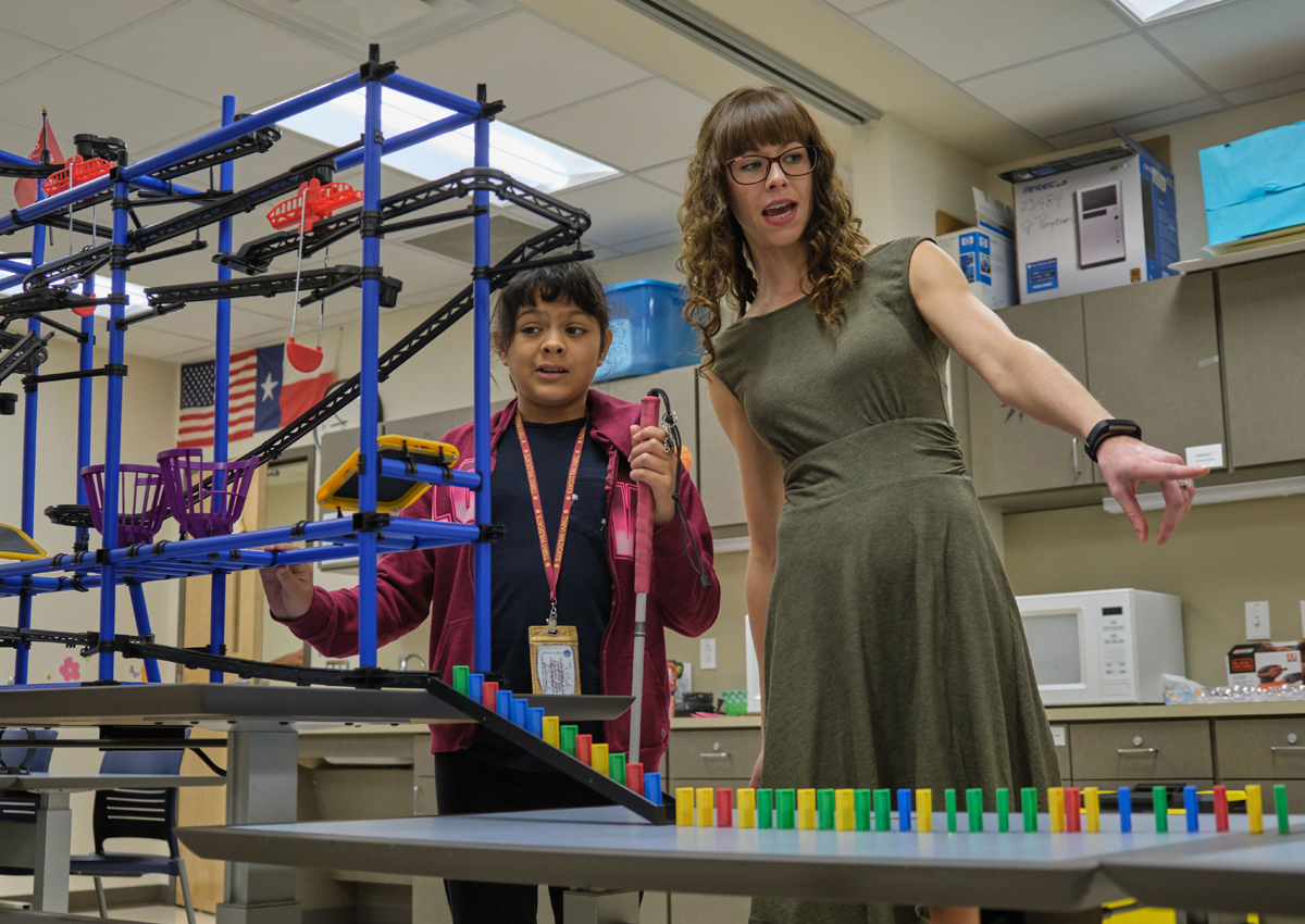 An adolescent with a white cane and an adult stand in front of a large, elaborate marble run contraption leading to a row of dominoes on a table.