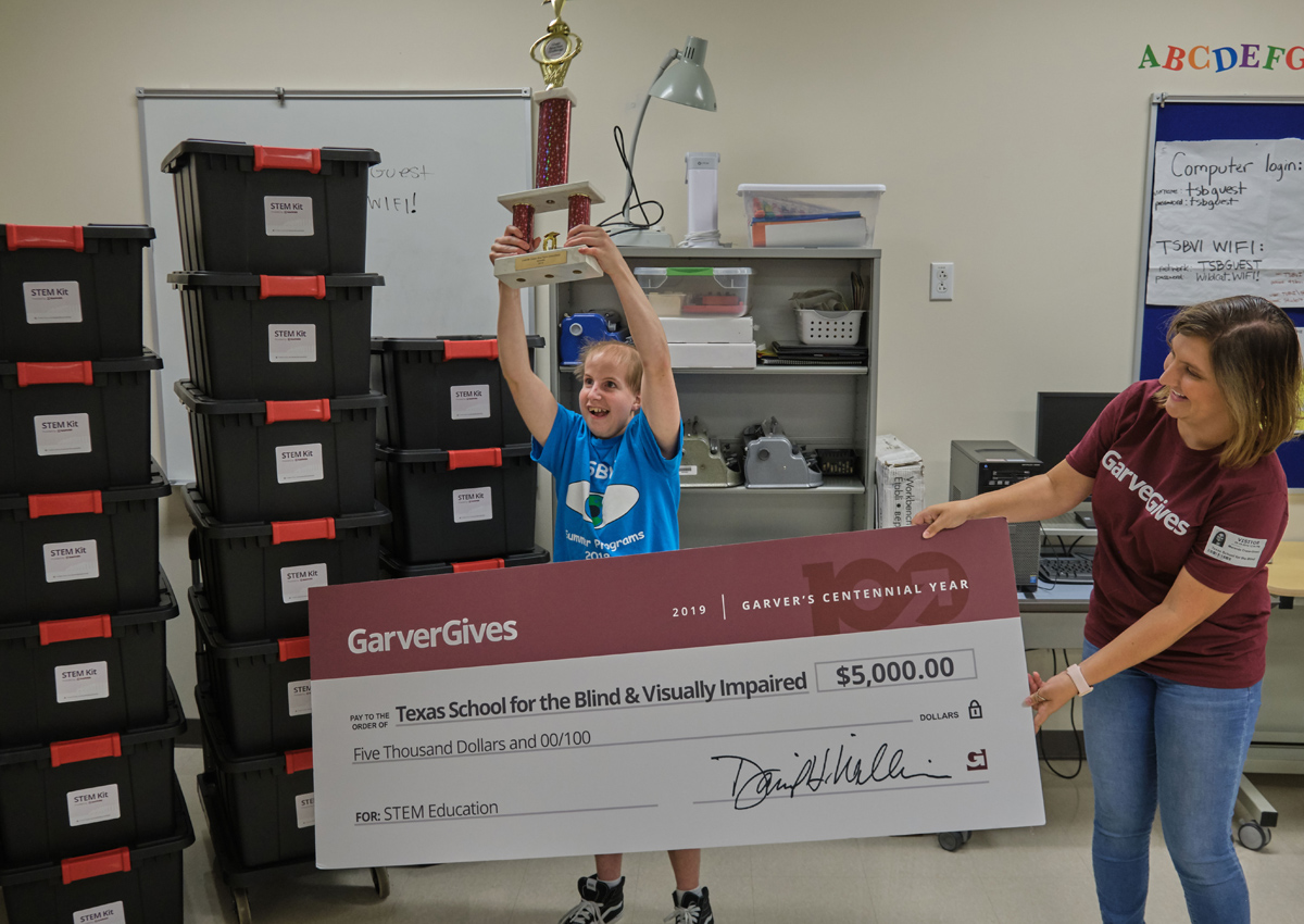 An adolescent holds up a trophy and stands in front of an adult holding a jumbo check from GarverGives for $5,000. About 20 large bins labeled 
