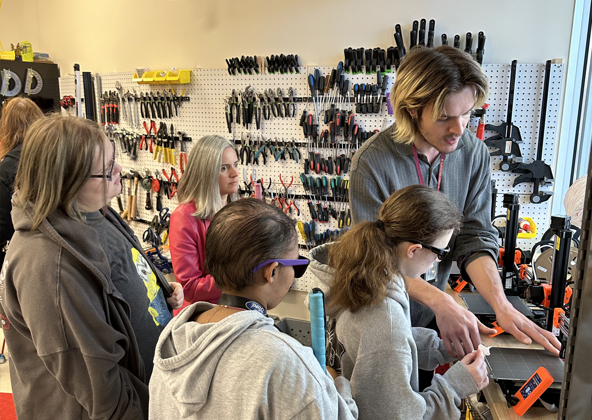 An adult demonstrates 3D printing to adolescents in a MakerSpace classroom with a 3D printing station and hundreds of tools organized on the wall.