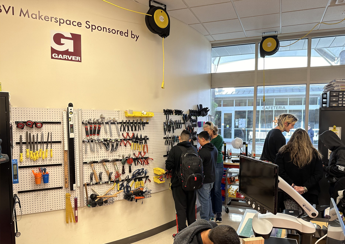 Adults and adolescents stand in front of a wall organized with various tools attached by peg board.