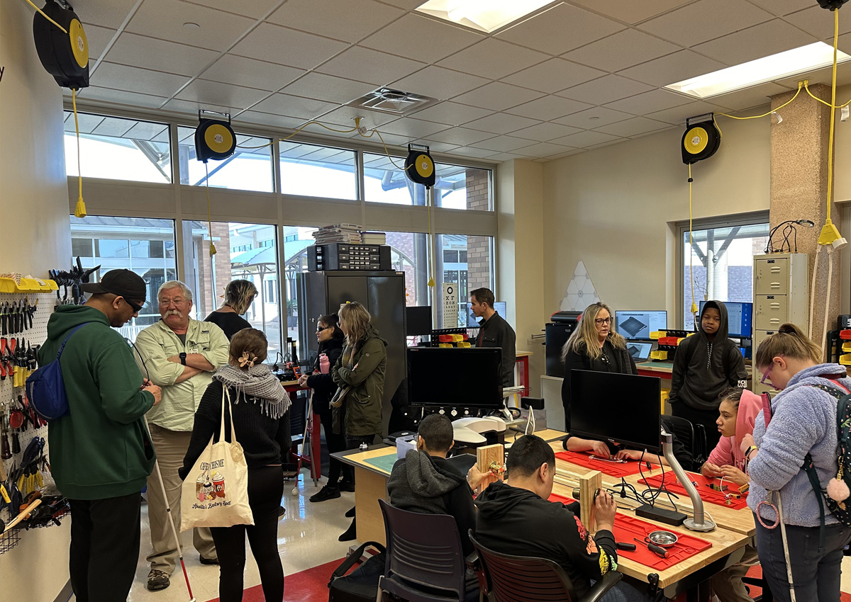 Adults and adolescents in a classroom equipped with organized tools attached to the wall, 3D printing stations, and wooden kits.