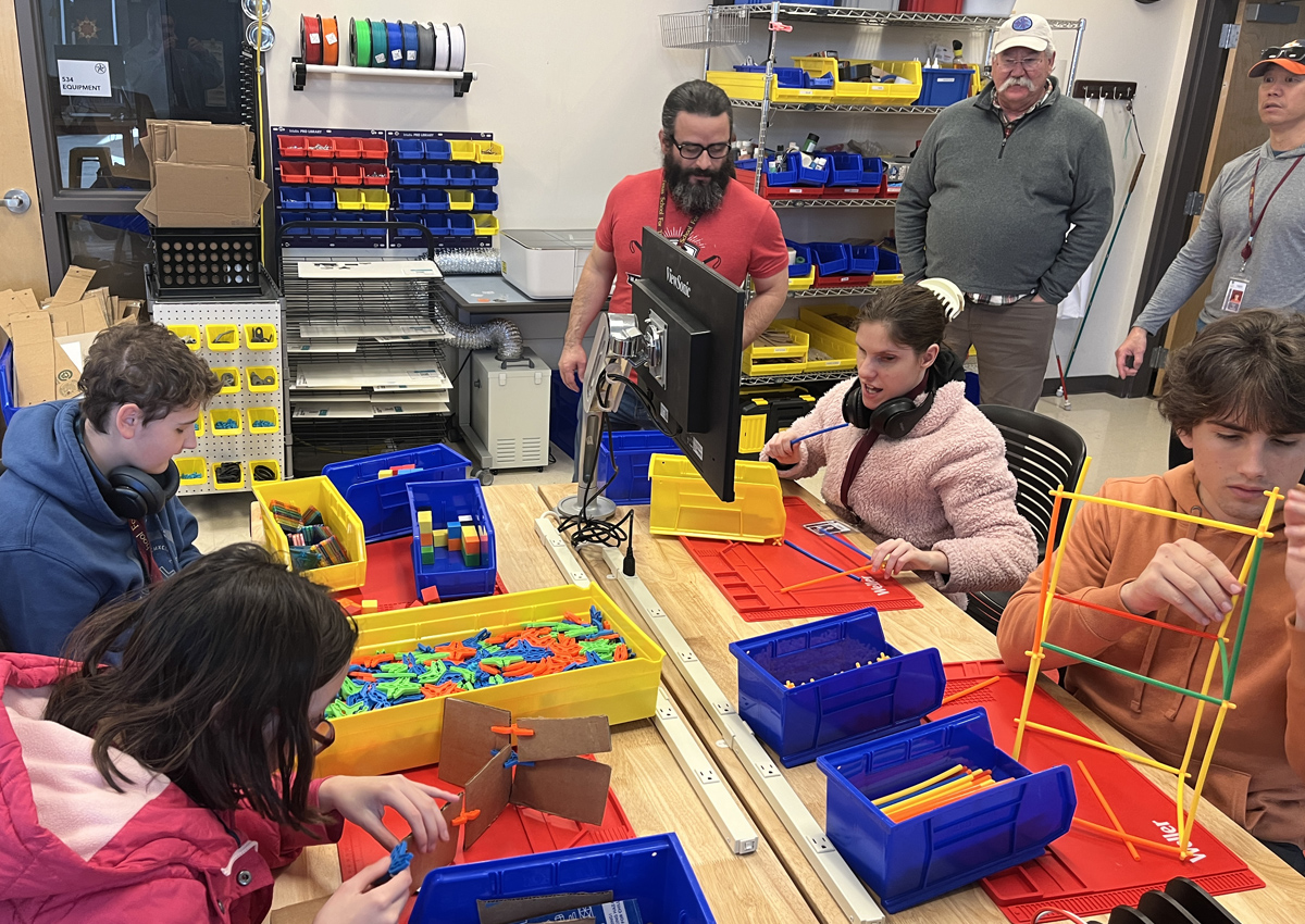 Four adolescents sit at a large work table with trays and bins. Some work building with straws and others build with cardboard and magnet tiles. Three adults stand nearby.