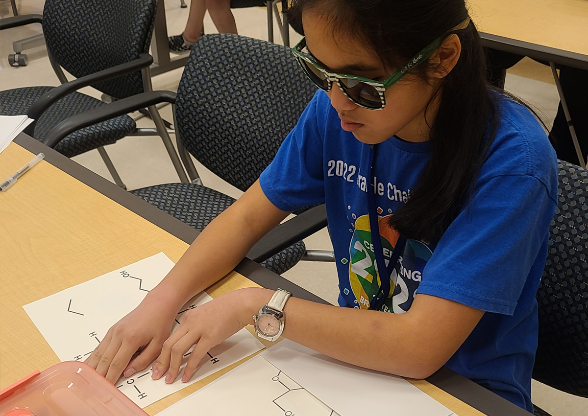 An adolescent explores tactile graphics of molecules on a piece of paper while seated at a table in a conference room.