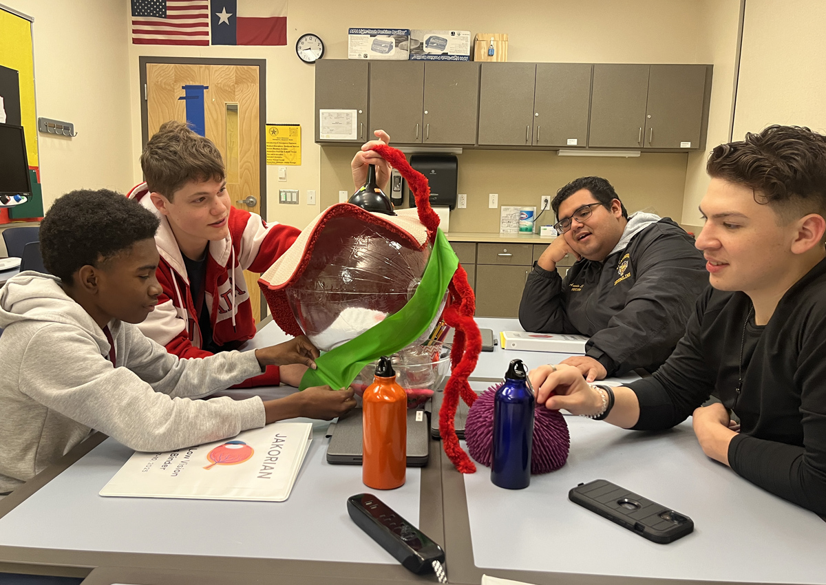 Four adolescents sit at a table and assemble the visual system made out of household objects (clear beachball, carpet, large pipe cleaners, squishy sensory ball, funnel, clear plastic bowl).