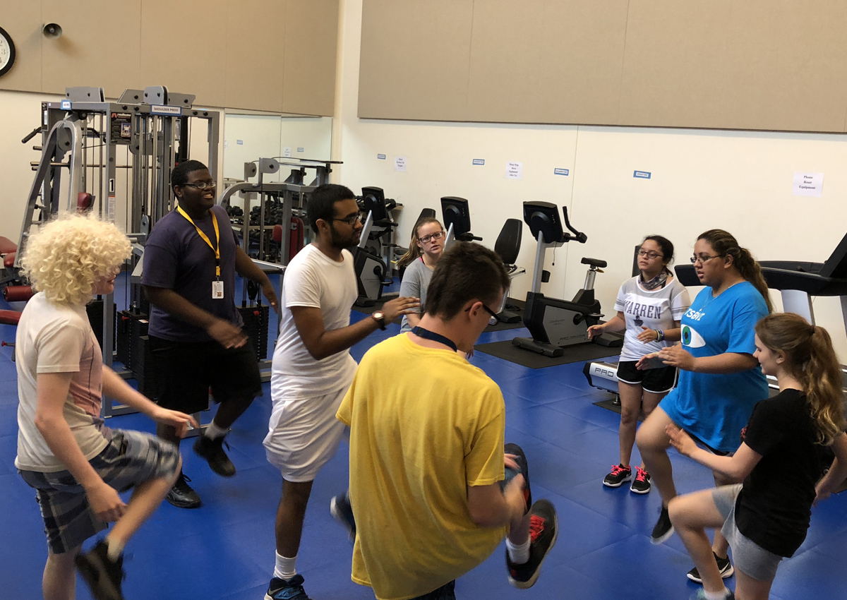 Eight adolescents kick up their knees while standing in place in a weight room.