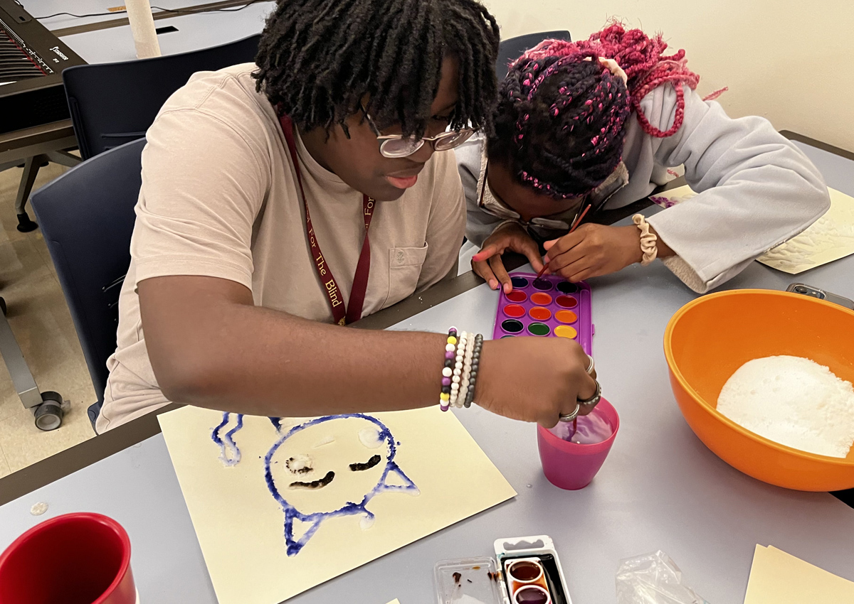 Dos adolescentes sentados en una mesa usan acuarelas para pintar dibujos, compartiendo materiales.