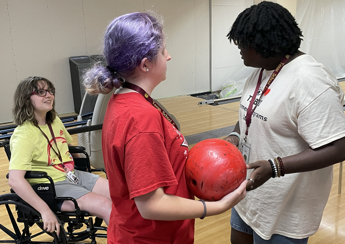 An adolescent hands a bowling ball to a peer while a third in a wheelchair looks on.