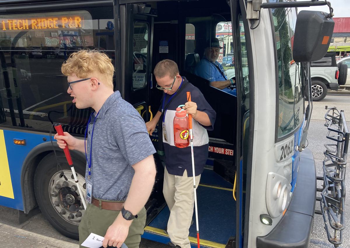 Two adolescents with white canes exit a city bus.