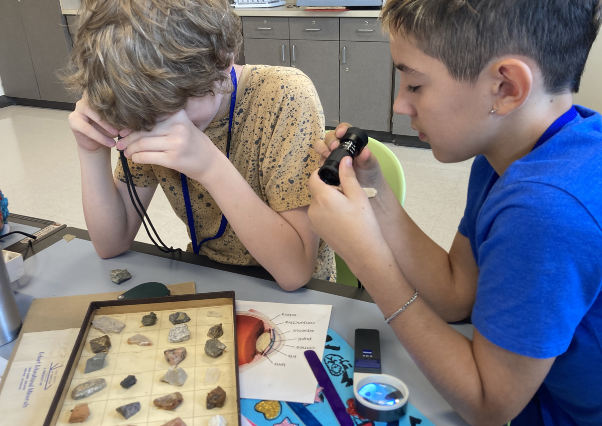 Two adolescents use telescopes and magnifiers to look at a tray of rocks and minerals. A diagram of the eye is on the table.