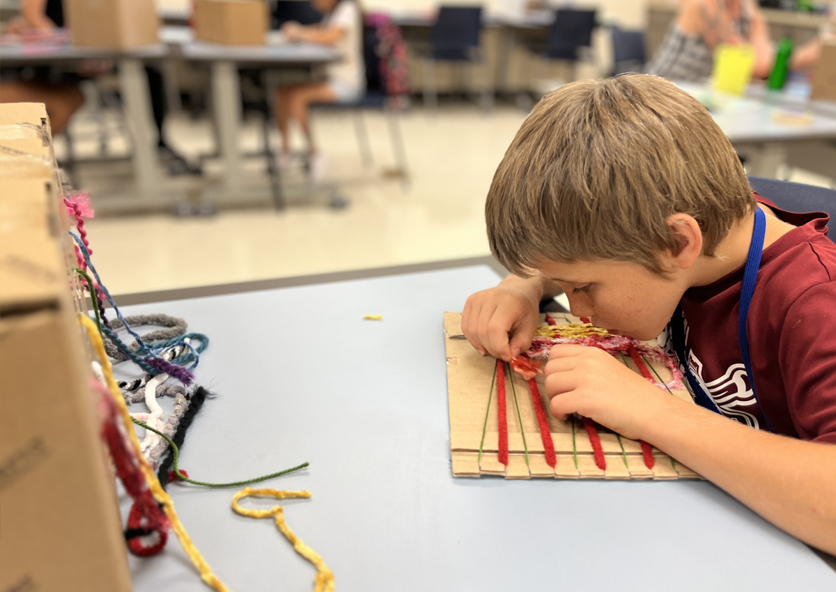 An elementary school aged student weaves yarn on a cardboard loom.