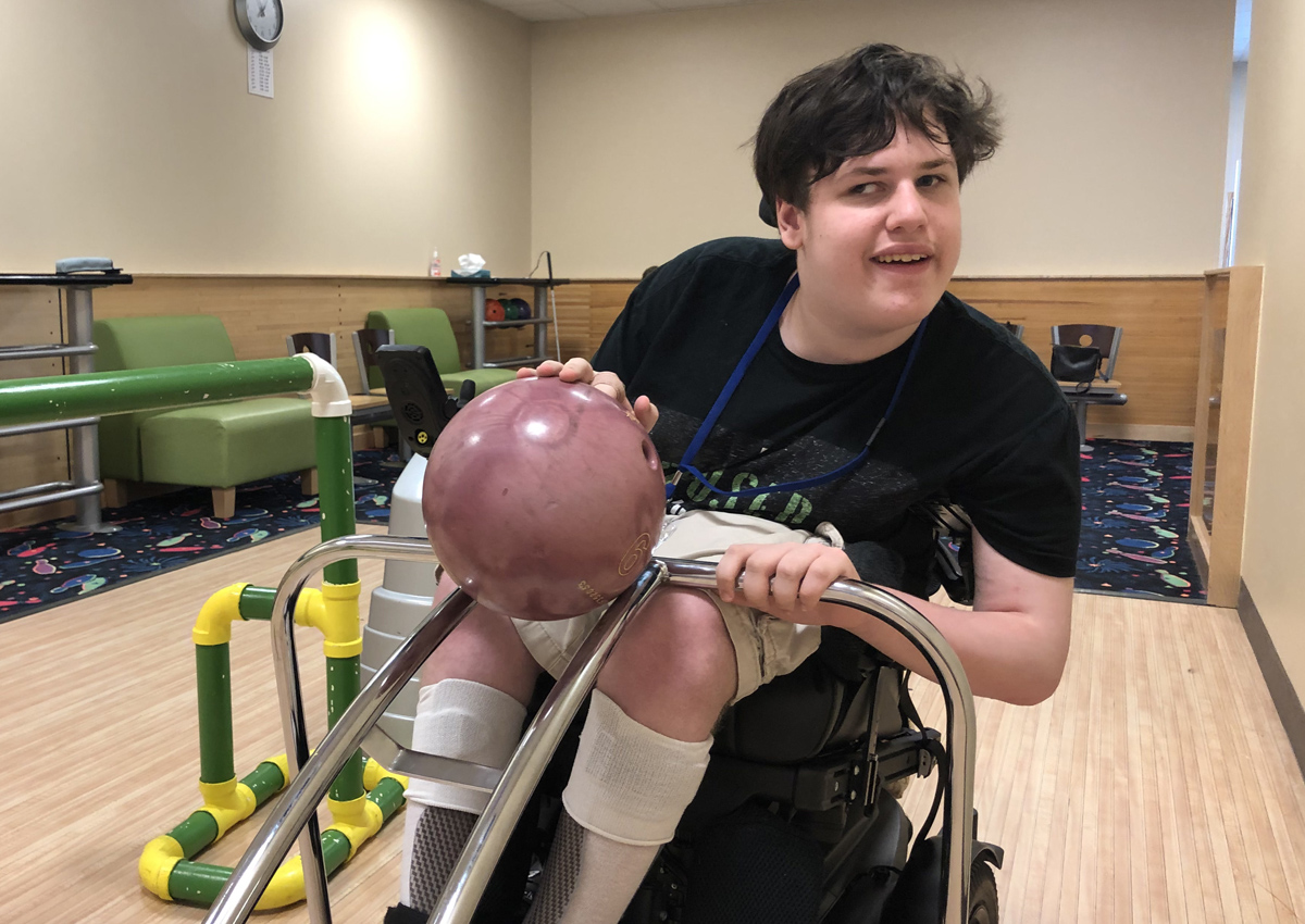 An adolescent in a wheelchair guides a bowling ball onto a ramp in a small bowling alley.