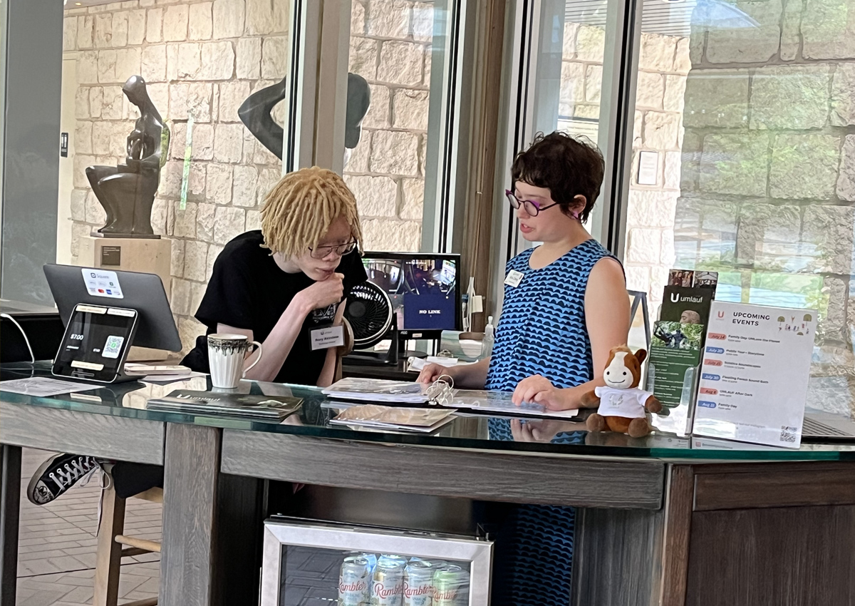 An adolescent and adult stand behind a museum desk in a lobby while looking in a binder together.