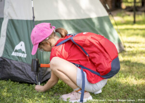 An adolescent wearing glasses and a backpack hammers in a tent spike in a state park. Photo credit: Courtesy Maegan Lanham, TPWD