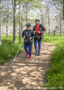 An adolescent using a straight cane and a second adolescent hike along a state park trail. Photo credit: Courtesy Maegan Lanham, TPWD