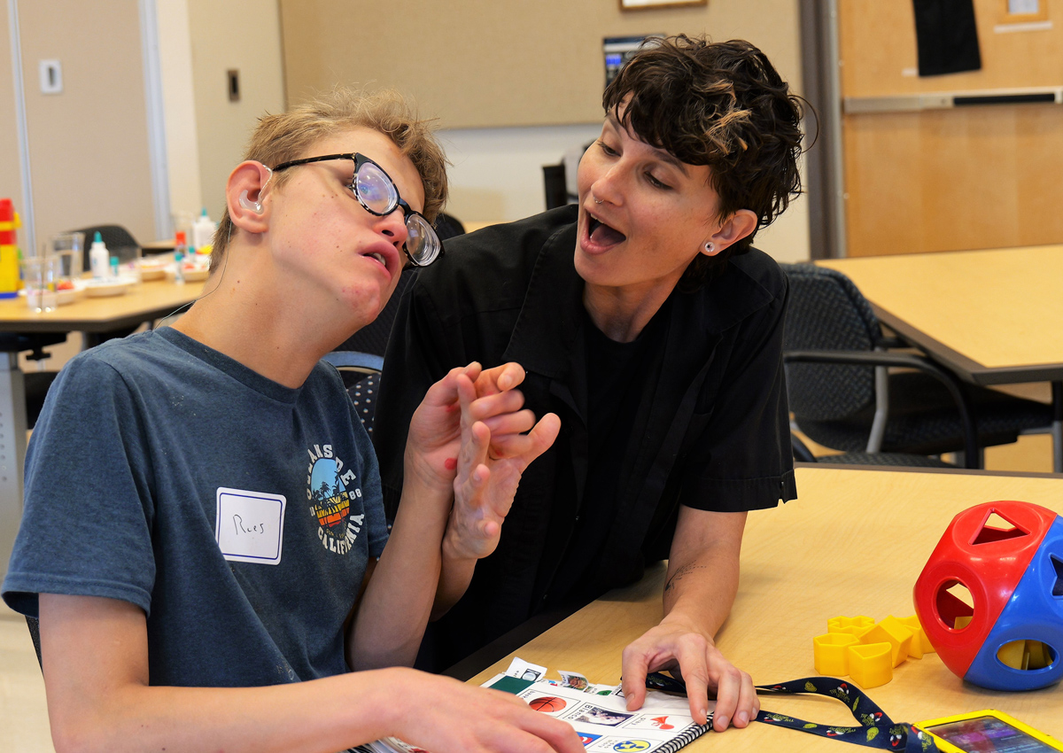 An adult uses tactile sign language with an adolescent wearing a hearing aid. On the table in front of them is a communication book with picture symbols.