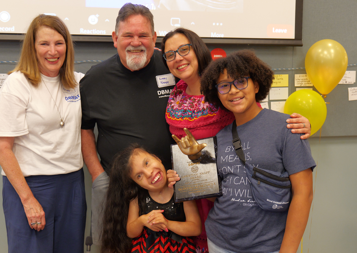 Two adults in DBMAT shirts pose with an adult and two adolescents while holding a plaque with a sculpture of a hand signing “I Love You” and the text: “DBMAT Christian Knapp Award, Great Motivator, Valentina Franco-Avila.”