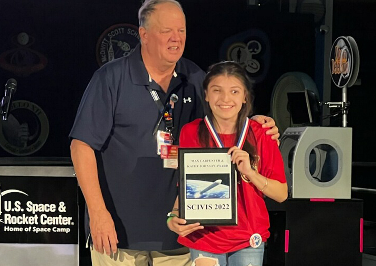 An adult presents an award to an adolescent with long, brown hair wearing a red Space Camp t-shirt, a medal, and holding the framed award.